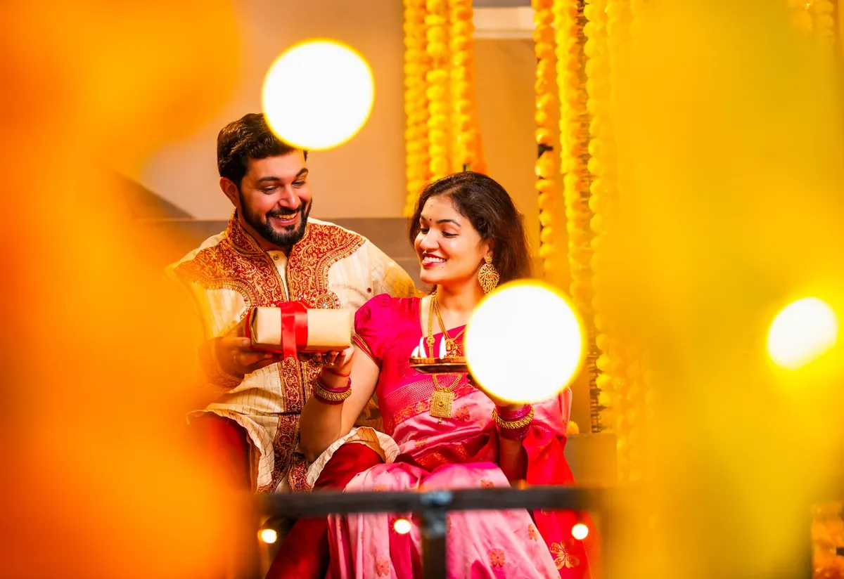A smiling man in a cream and red embroidered kurta holds a wrapped gift beside a smiling woman in a pink sari who holds a small plate with lit lamps, framed by marigold garlands and glowing lights