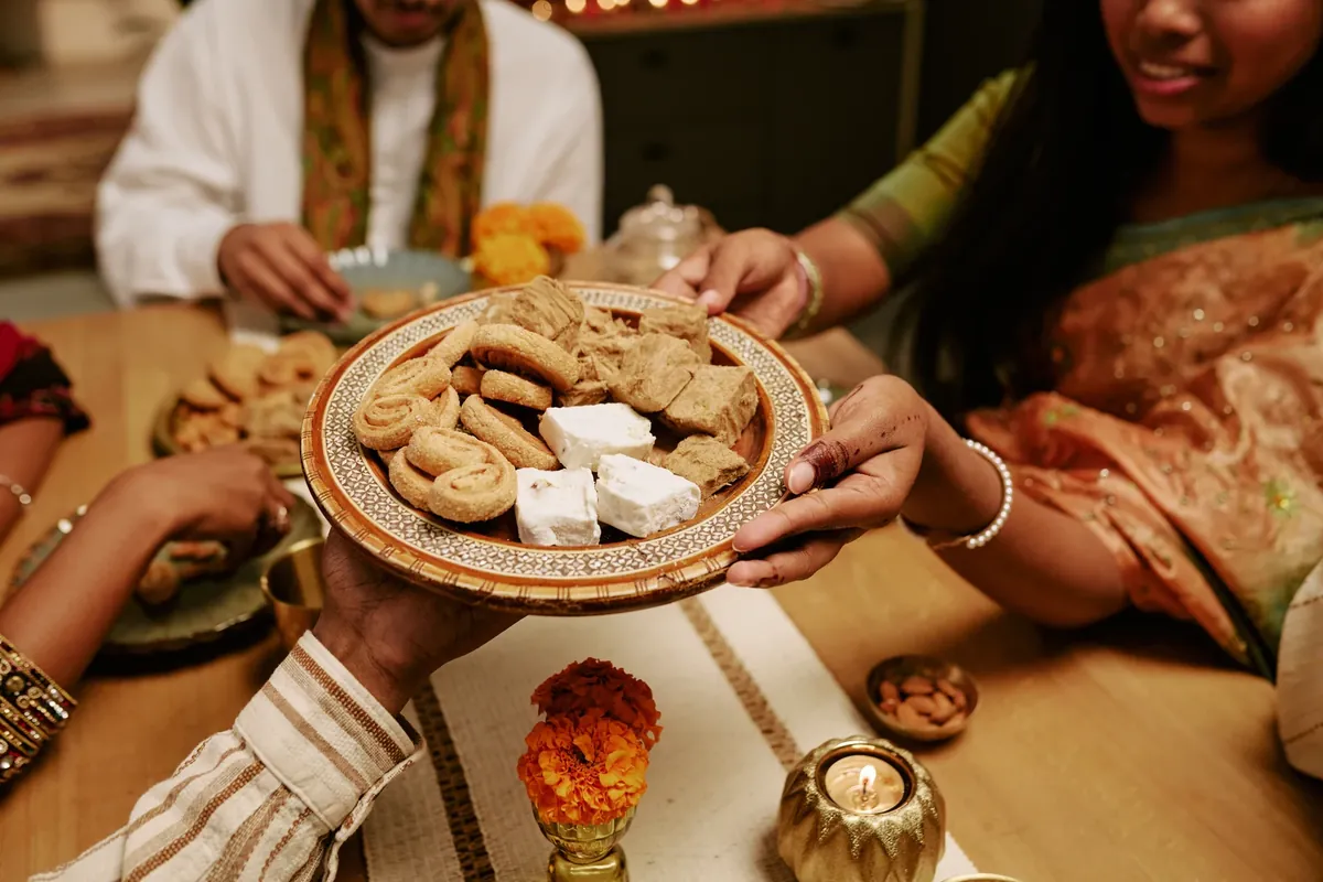 Hands passing a decorated plate of Indian sweets across a table set with marigolds, a small lamp and almonds