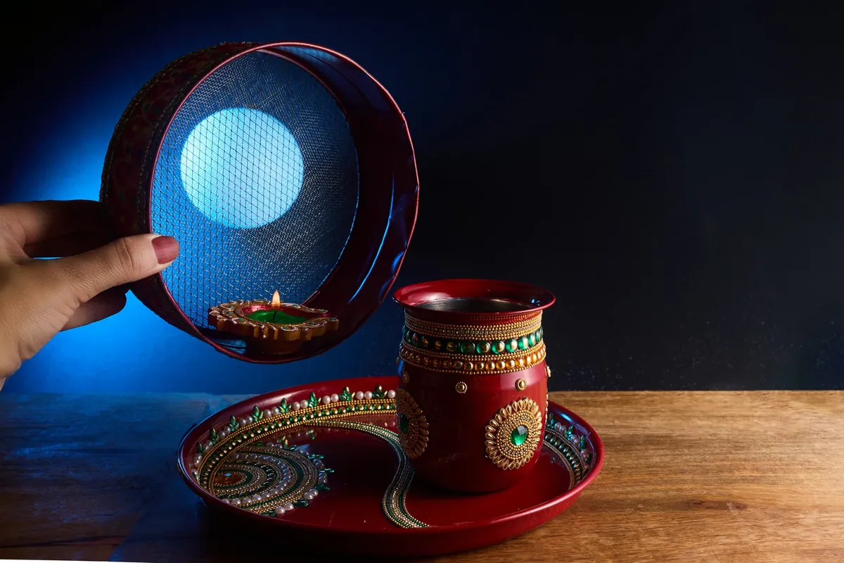 A decorated red karwa pot and thali on a wooden table, with a hand holding a sieve in front of a glowing disc and a small lit lamp
