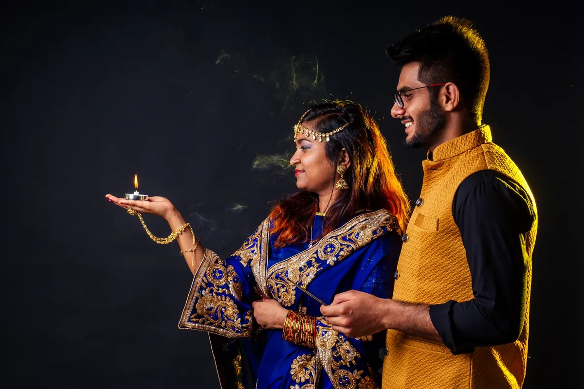 A woman in a blue and gold sari with a gold head ornament holds a small lit tealight in her palm while a smiling man in a mustard waistcoat stands behind her, against a dark background