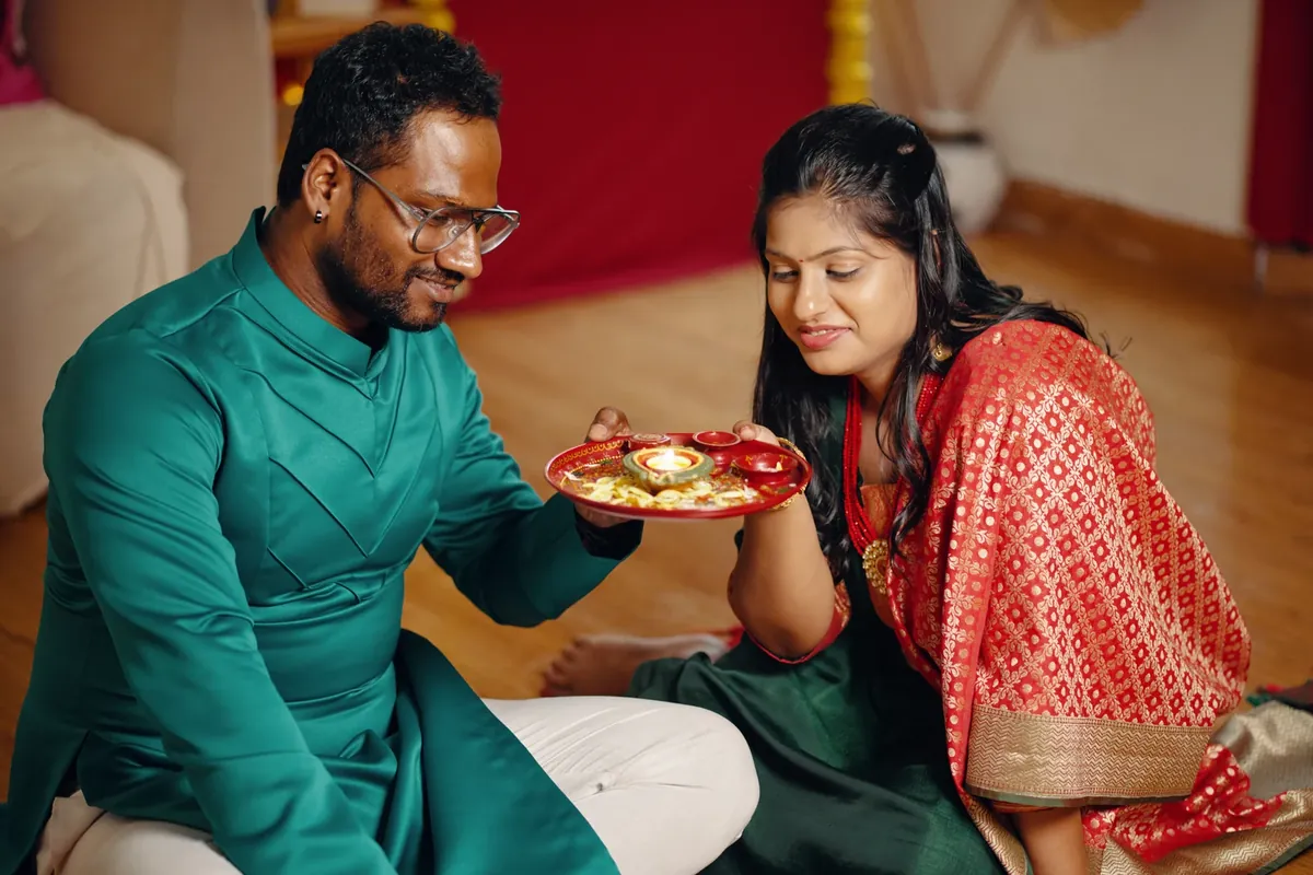 A man in a green kurta holds a small red plate with a lit diya while a woman in a red patterned sari looks at it