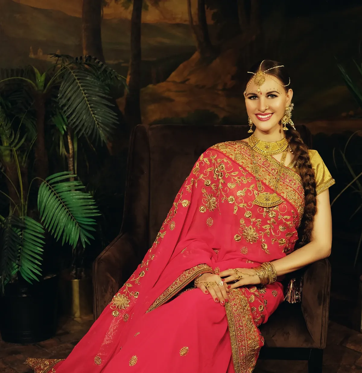 A woman in a pink saree with gold embroidery, gold jewellery and a maang tikka sitting in an armchair