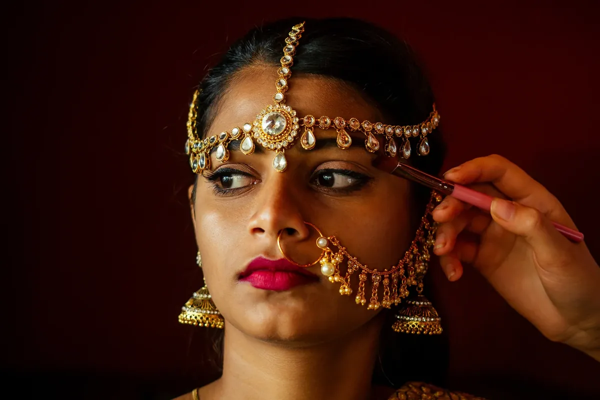 A makeup artist brushes eye makeup on a woman wearing a gold maang tikka, pearl nose ring and jhumkas