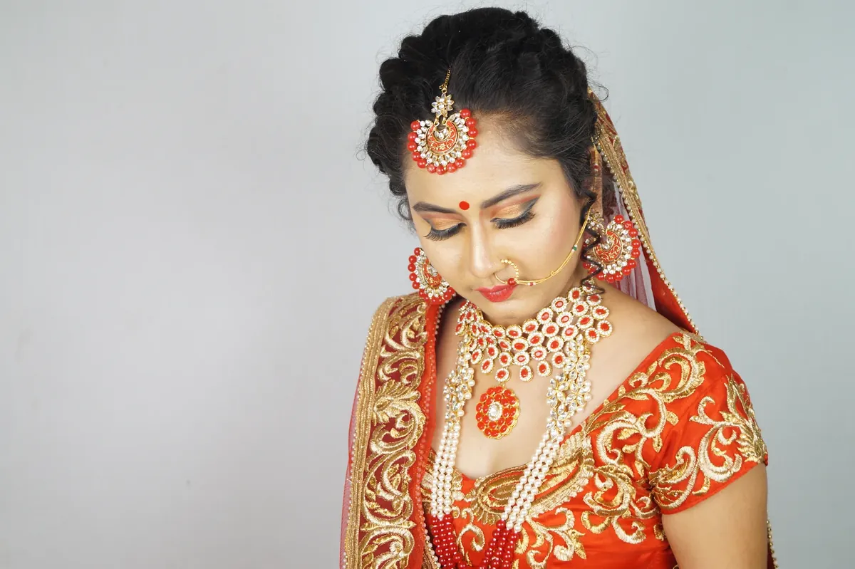 A woman in red and gold wearing a bindi, maang tikka, nose ring, earrings and a layered red and pearl necklace