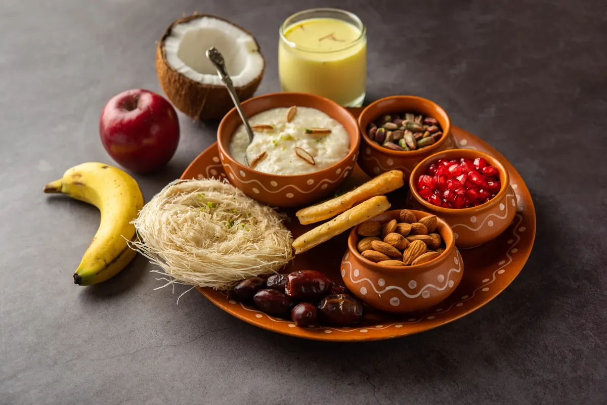 A terracotta-coloured plate with small bowls of rice pudding, pistachios, pomegranate seeds and almonds, plus dates, a nest of vermicelli sweet, two fried sticks, a banana, an apple, half a coconut and a glass of milk