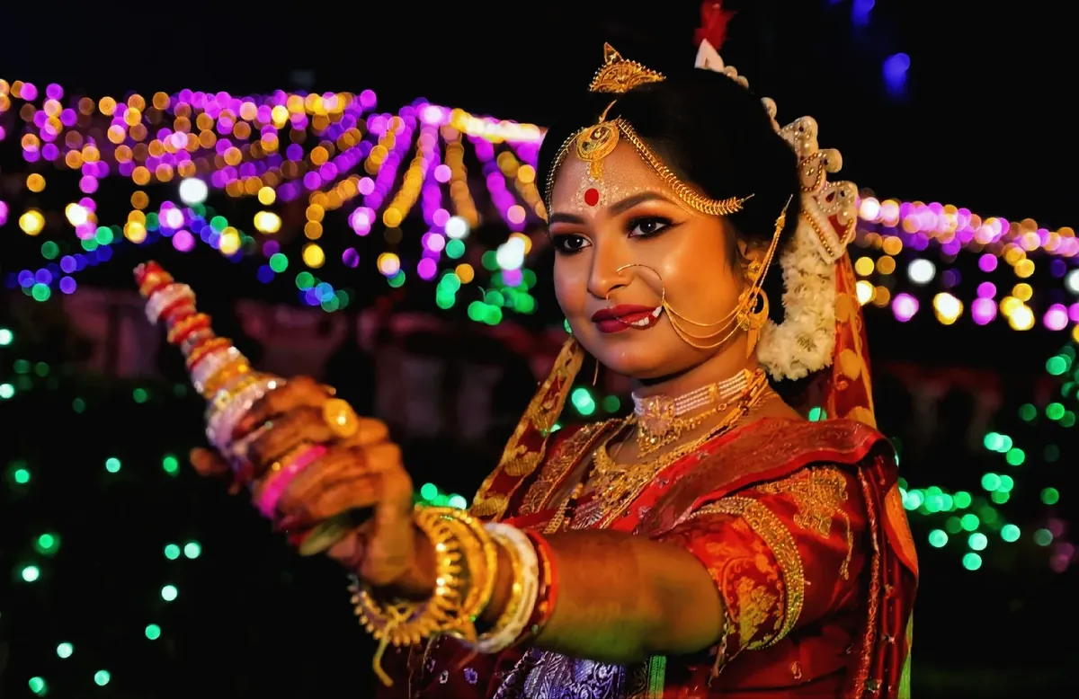 Bengali-style bride in a red saree with a nose ring, gold necklaces and white and red bangles, lit by festival lights