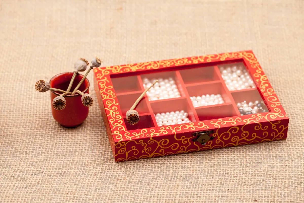 Red painted jewellery box with a clear lid and pearl-filled compartments, beside a small red cup of dried poppy pods