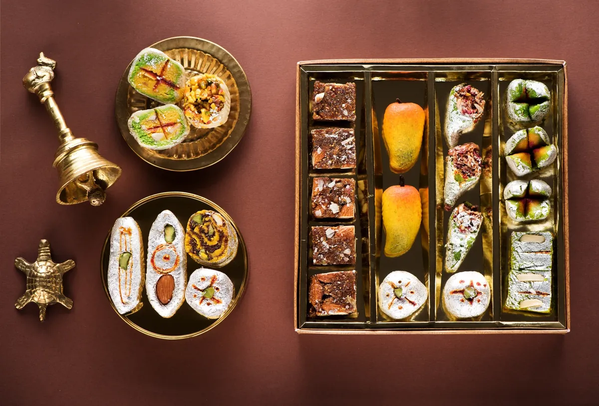 Brass plate of sweets and a box of assorted mithai in rows, with a small brass bell and tortoise beside them