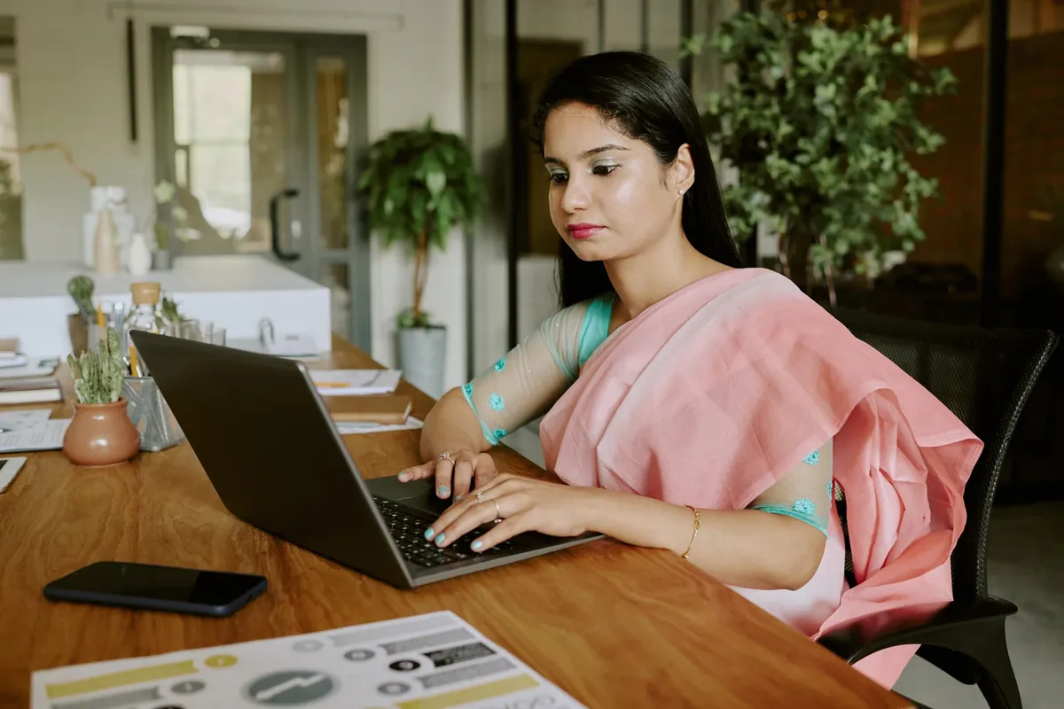 A woman in a pink saree typing on a laptop at a wooden desk with a phone and printed papers beside her