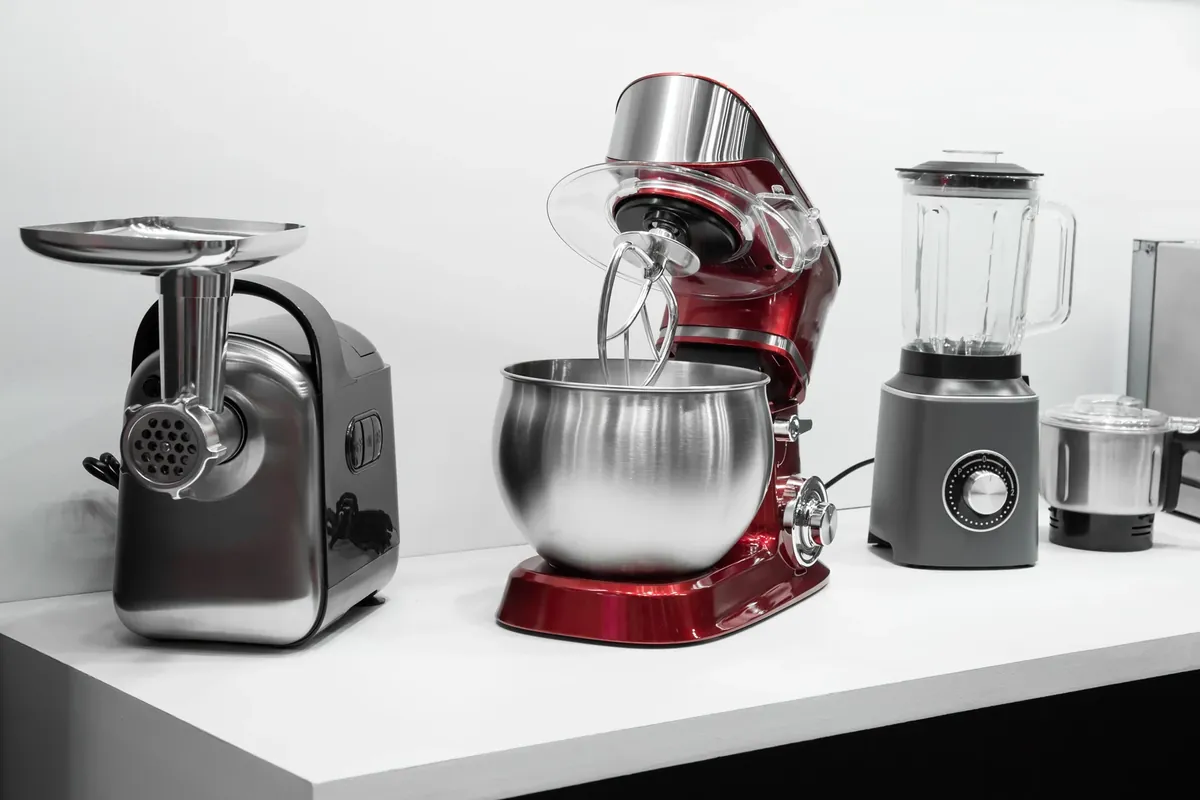 A red stand mixer with a steel bowl, a blender and a mincer lined up on a white counter
