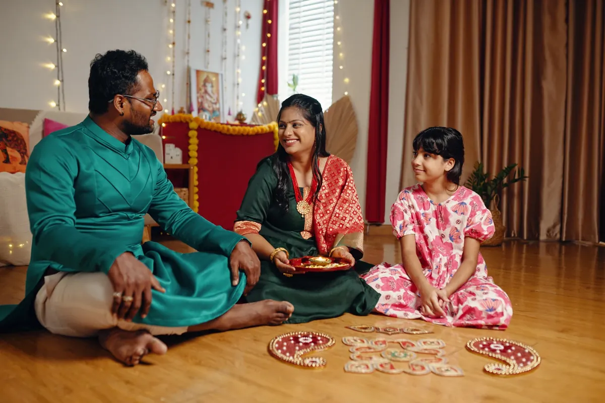 A family sits on a wooden floor in a room with fairy lights and a Ganesha picture, with decorative rangoli pieces laid out in front of them while the woman holds a lit diya on a plate