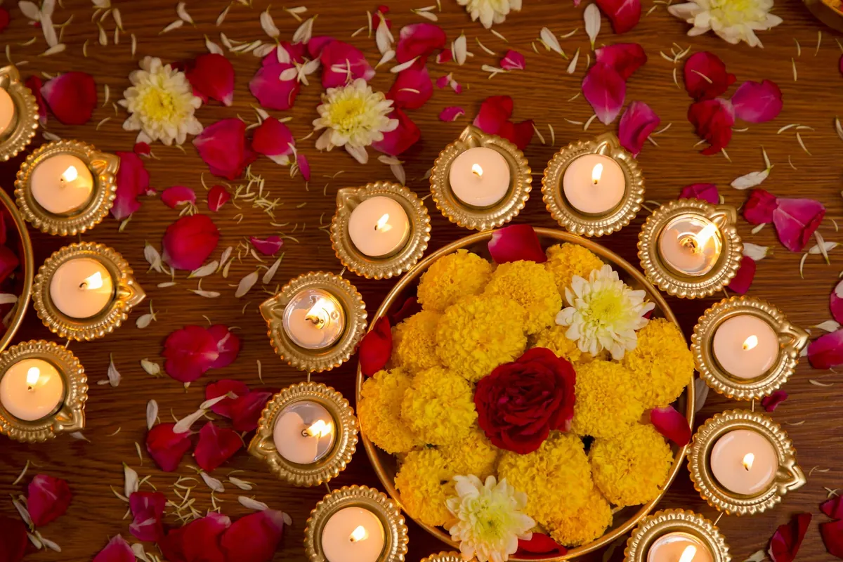 Brass diyas with lit tealights arranged around a brass bowl of yellow marigolds and a red rose, with red and white petals scattered on a wooden floor