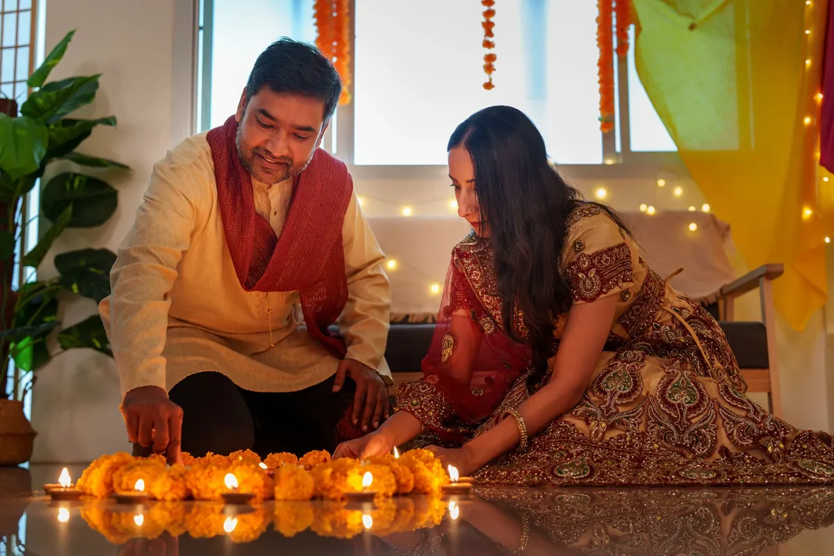 A man in a cream kurta and a woman in a maroon outfit set lit diyas along a row of marigolds on a living-room floor