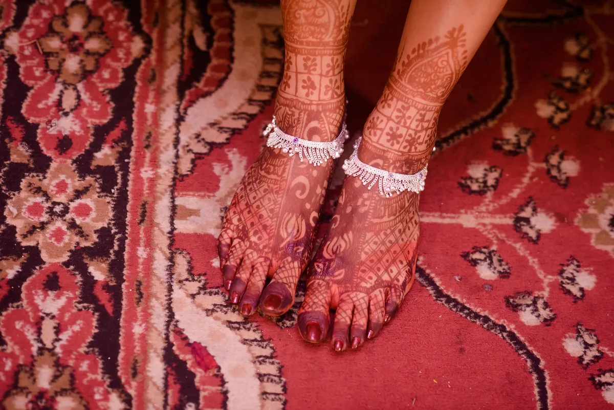 Feet decorated with henna wearing silver payal anklets on a red patterned rug