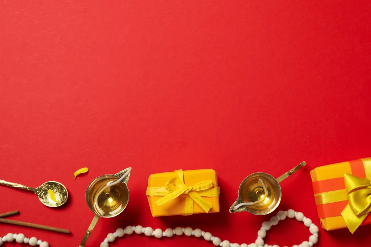 Brass diyas, a spoon, a white bead mala and yellow gift boxes tied with ribbon, laid out on a red background