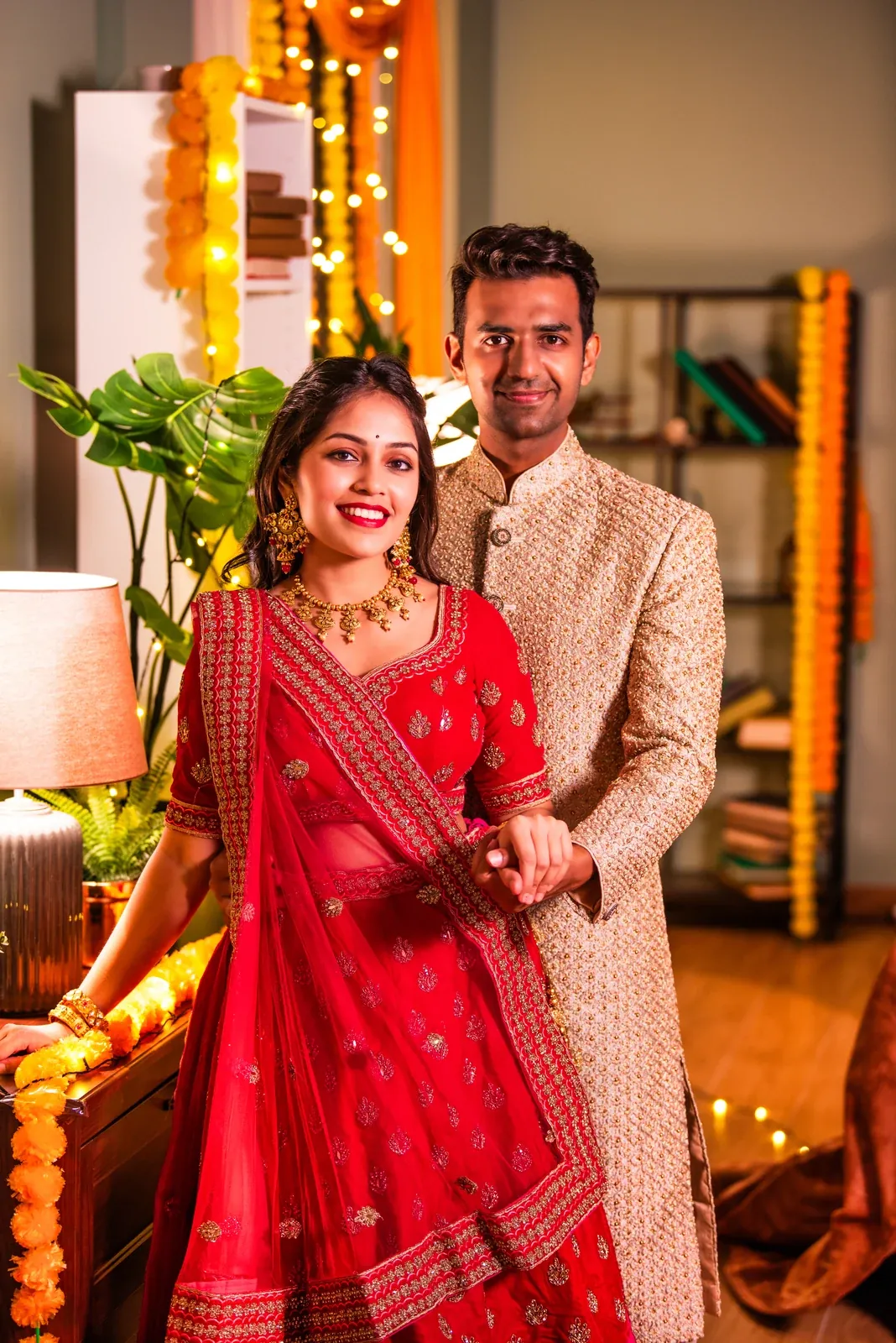A smiling couple in wedding clothes, she in a red lehenga and dupatta, he in a cream sherwani, at a decorated home