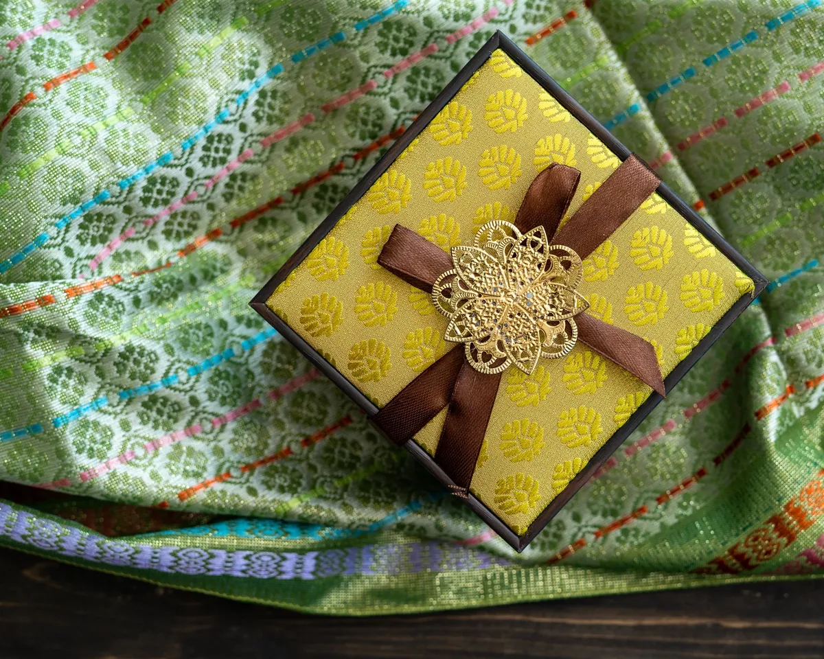 A gold-patterned gift box tied with a brown ribbon and a gold filigree ornament on green woven fabric