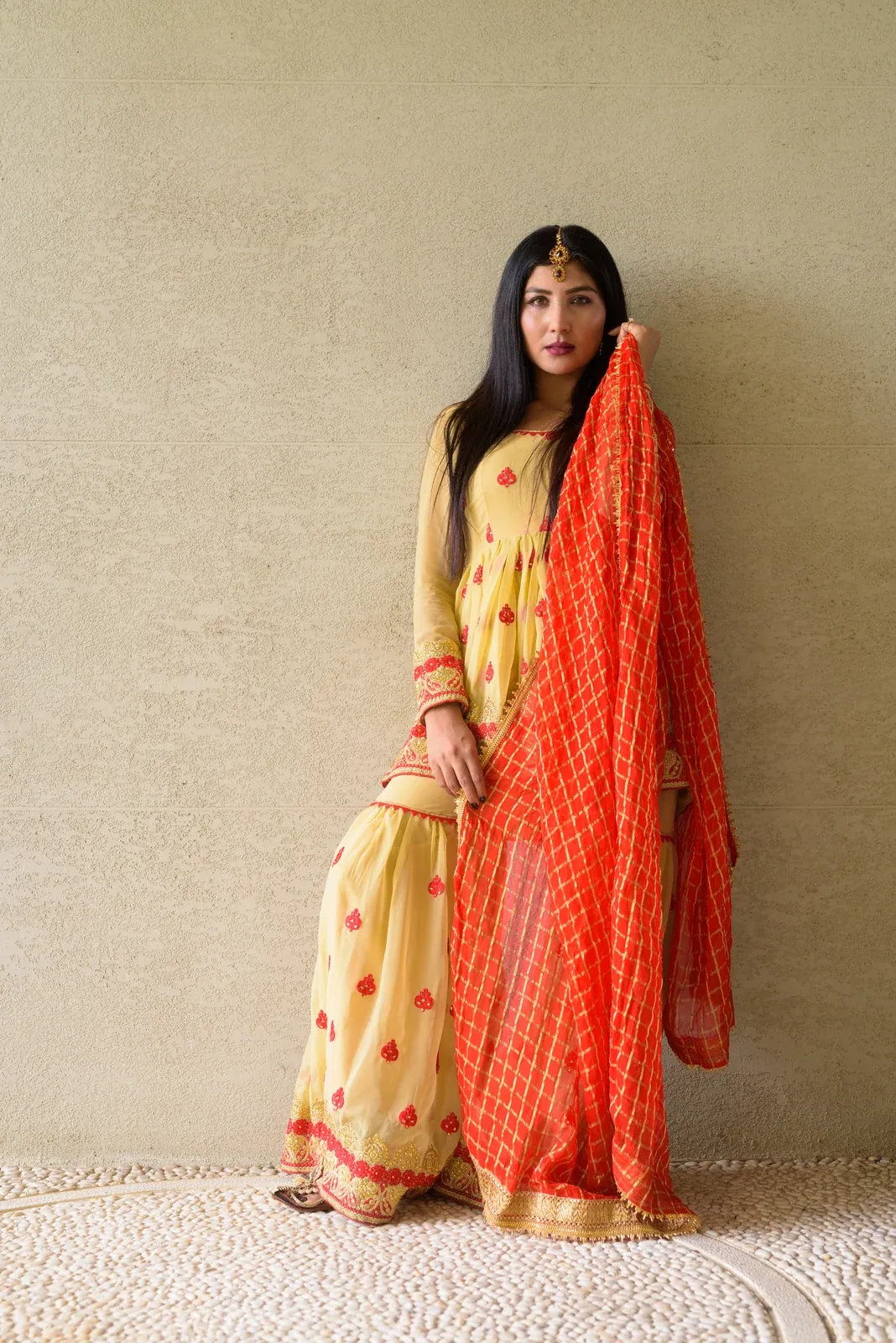 A woman in a cream full-sleeved suit with red motifs holds an orange checked dupatta draped from her shoulder