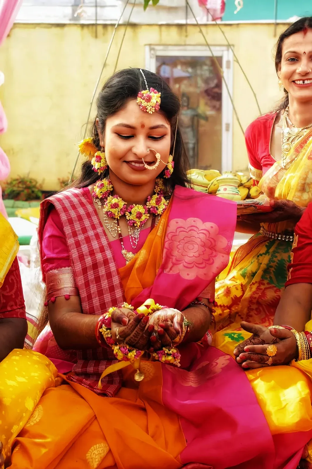 A smiling woman in a pink and orange saree with yellow flower jewellery and mehndi on her hands at a wedding ceremony