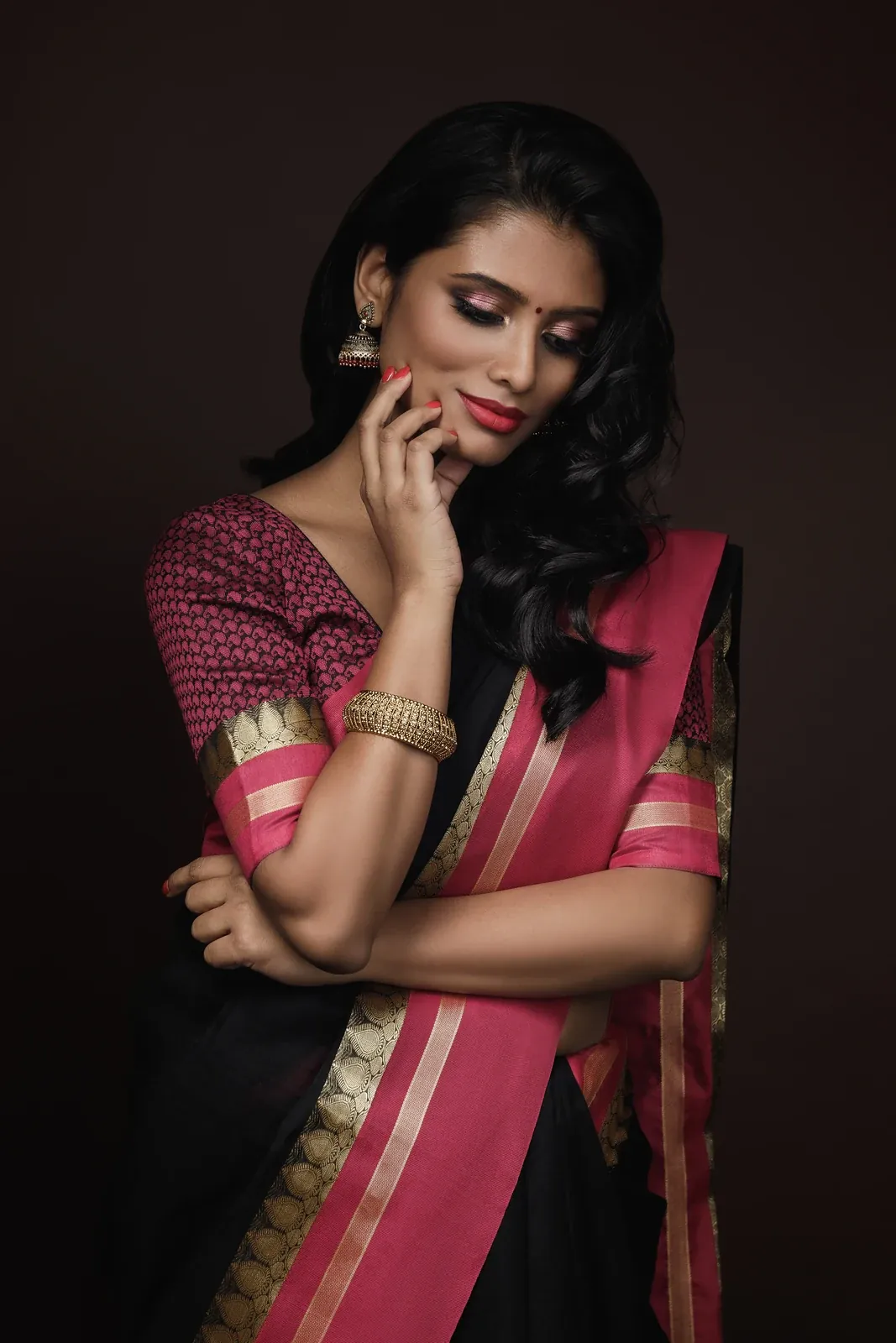 A woman in a black and pink silk saree with a wide gold zari border, one hand at her chin, against a dark background