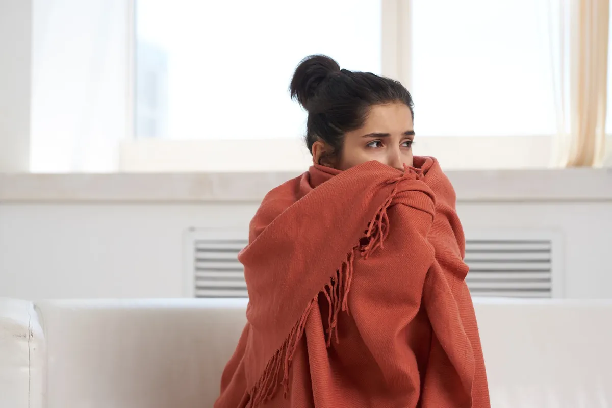 A woman wrapped in a rust-orange shawl holds it close against the cold, seated on a white sofa by a window