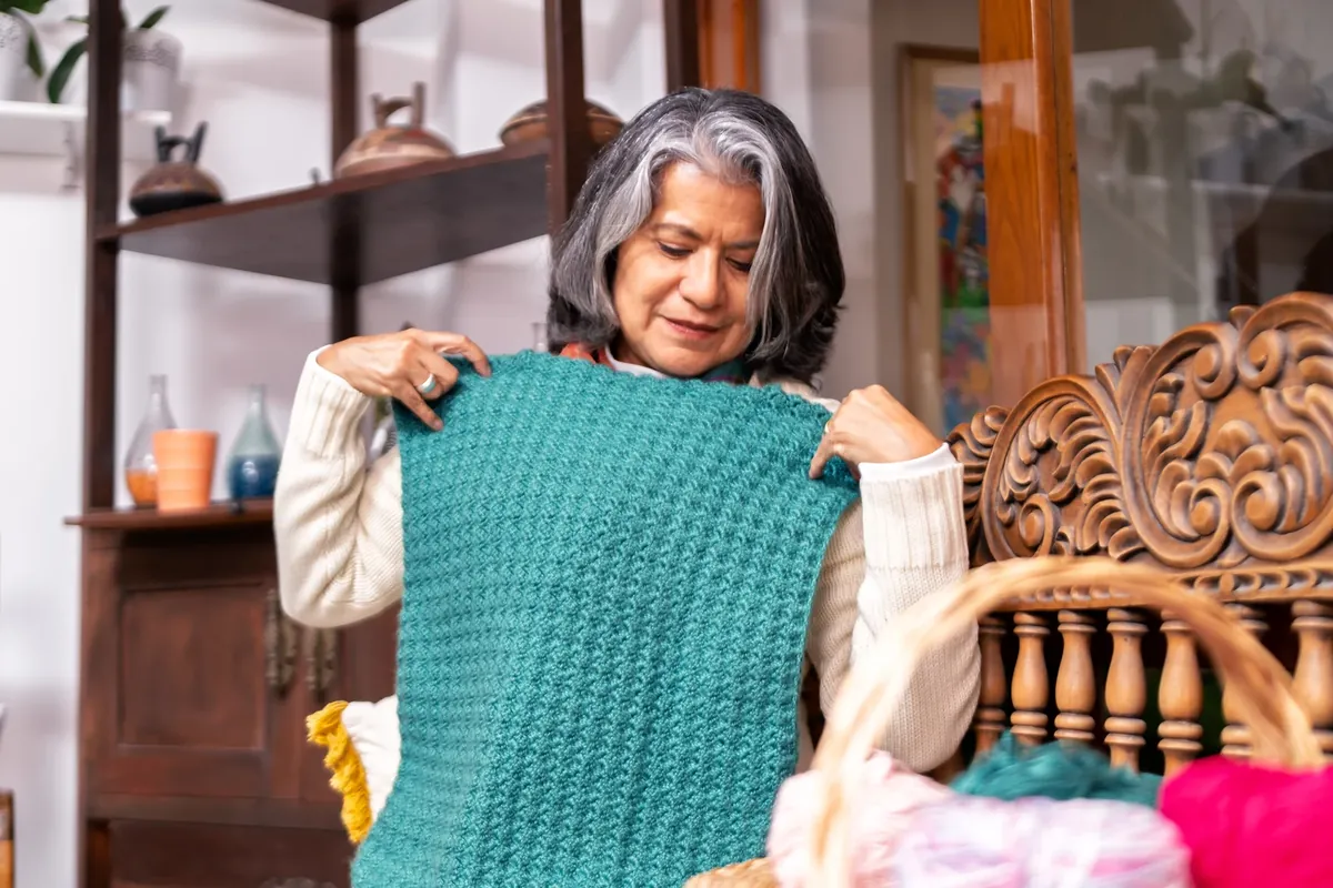 A woman with grey hair holding up a teal knitted piece to look at it, with balls of wool in a basket in front of her