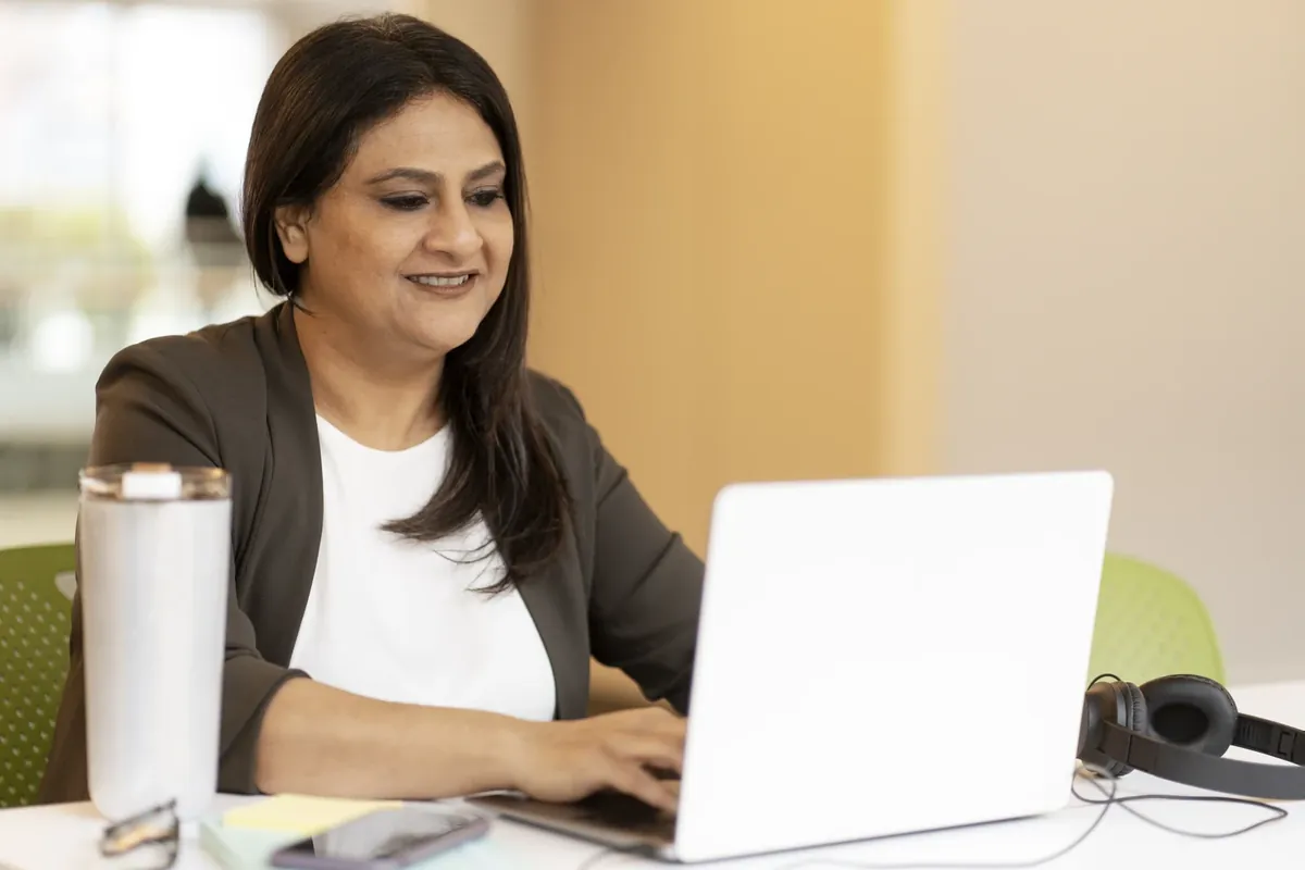 A smiling woman in a dark blazer over a white top types on a laptop at a desk in a bright office